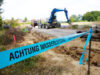 Absperrung "Achtung Wasserleitung", dahinter eine Baustelle mit offenem Boden. Es ist ein Bagger und Rohrleitungen zu sehen.